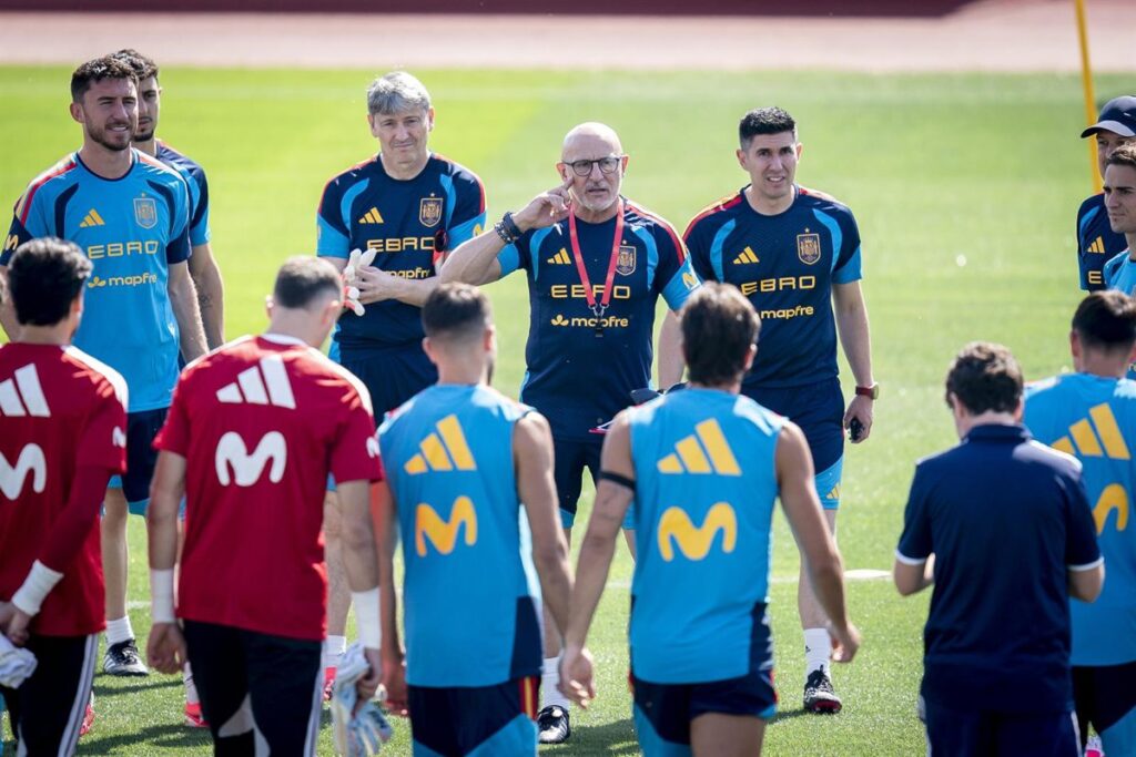 Lamine Yamal en entrenamiento con España 2026, sonriendo junto a Rodri y Gavi en un campo de fútbol con la bandera nacional